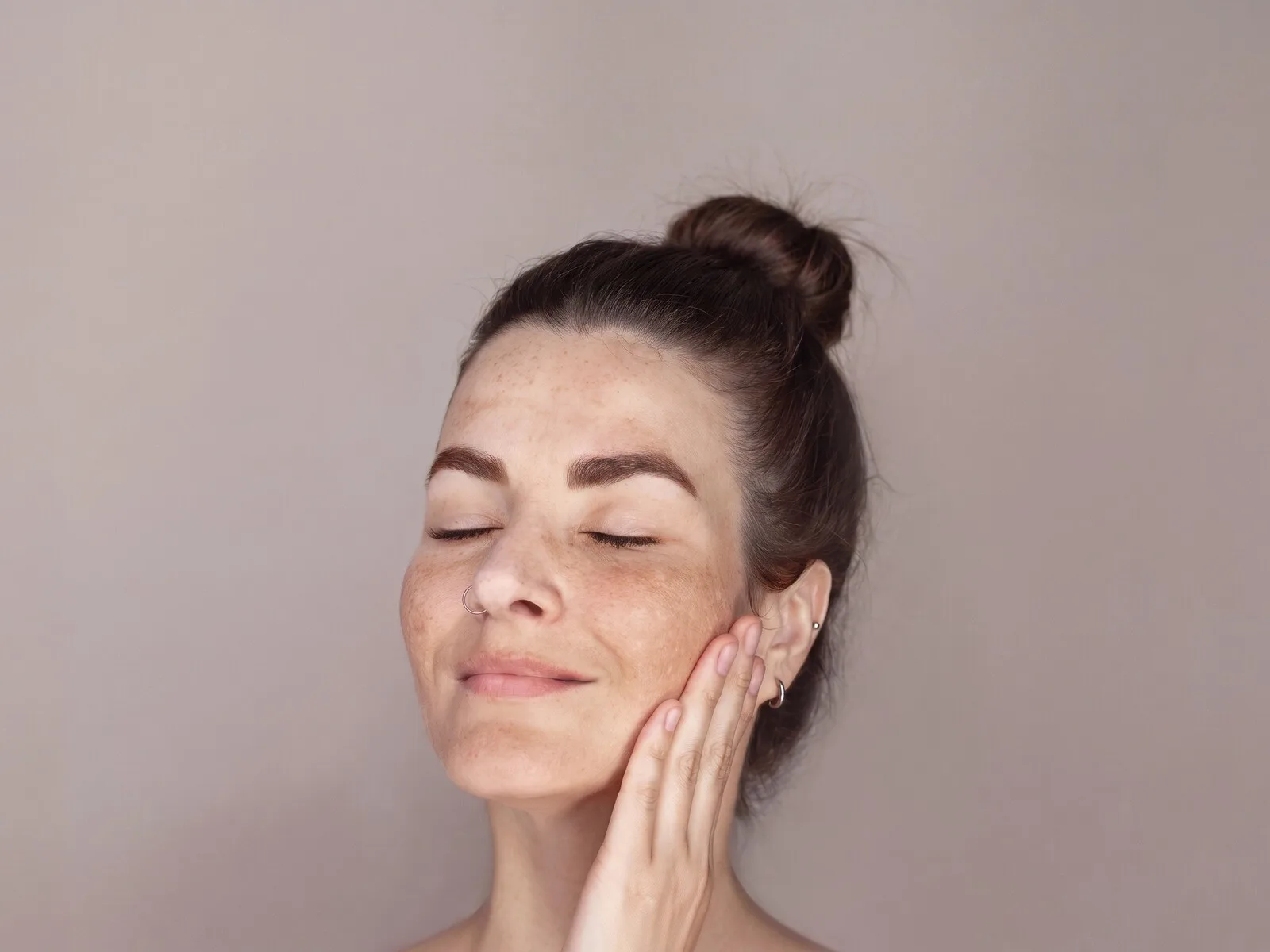 A close-up of a woman with her eyes closed and her fingertips resting against her cheek, freckling and soft brown melasma patches spread across both cheeks, the bridge of her nose and her upper lip