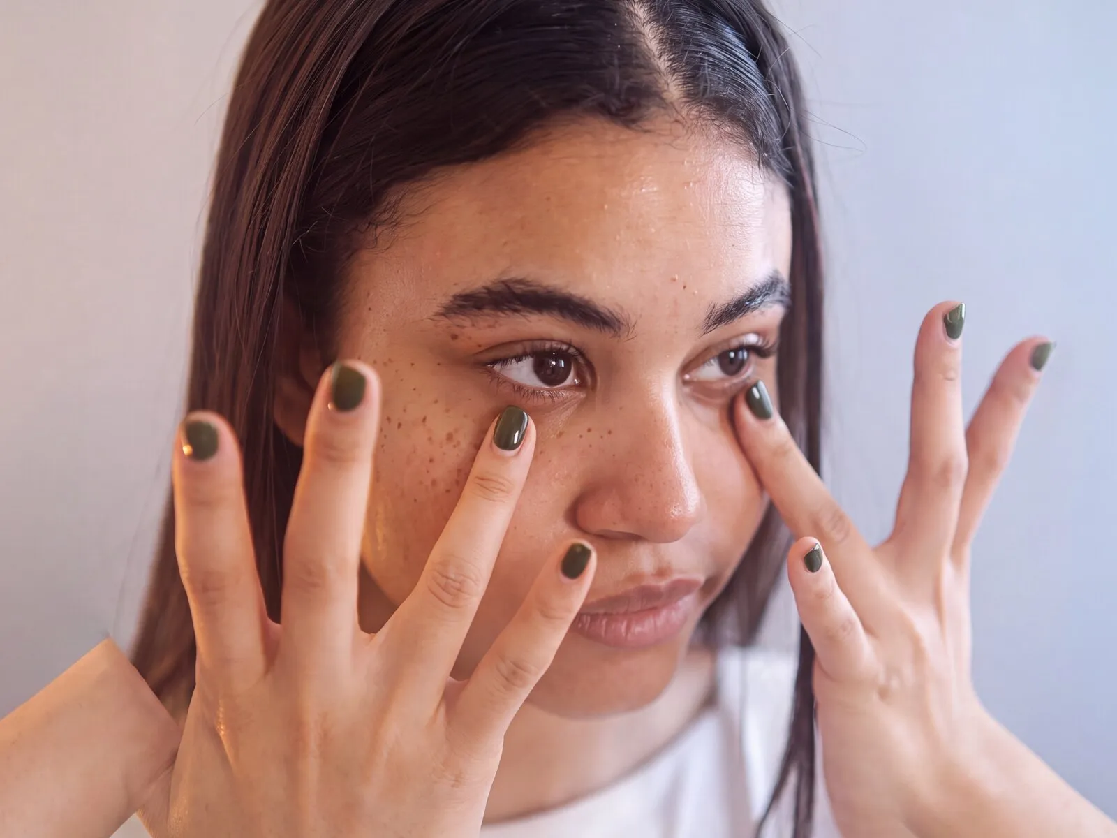 A woman with a fingertip resting below each eye, both hands raised, soft brown-violet periorbital shadowing under both lower lids