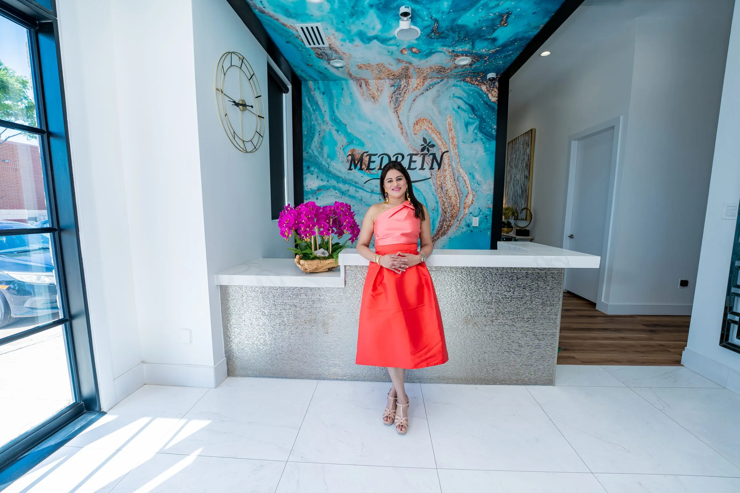 Dr. Haritha Mikkilineni, MD, standing at the reception desk of the Medrein Health clinic in Southlake, TX, beneath the practice’s blue marble mural and wordmark
