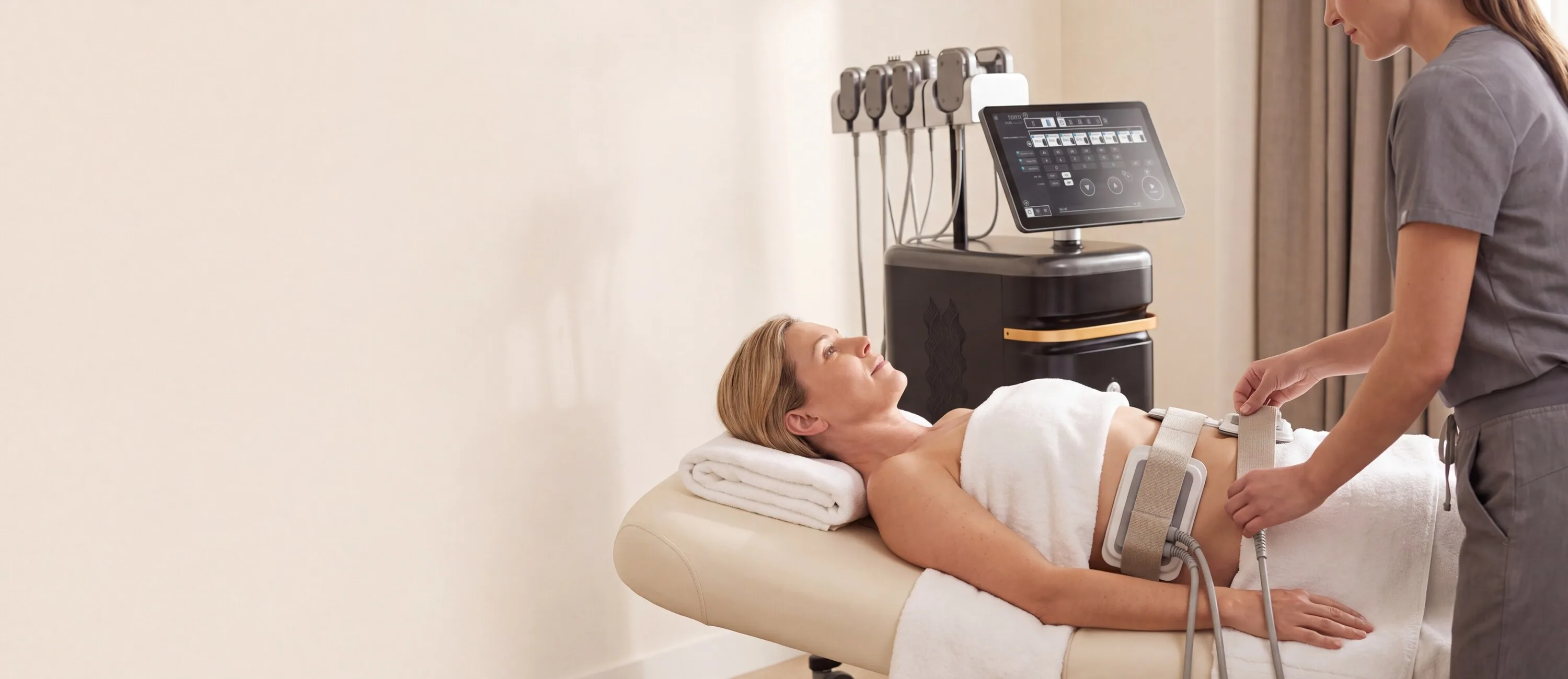 A draped patient on a treatment couch with body-contouring applicator pads belted across her abdomen while a clinician adjusts the strap, the MNML Tone console and its docked handpieces behind them