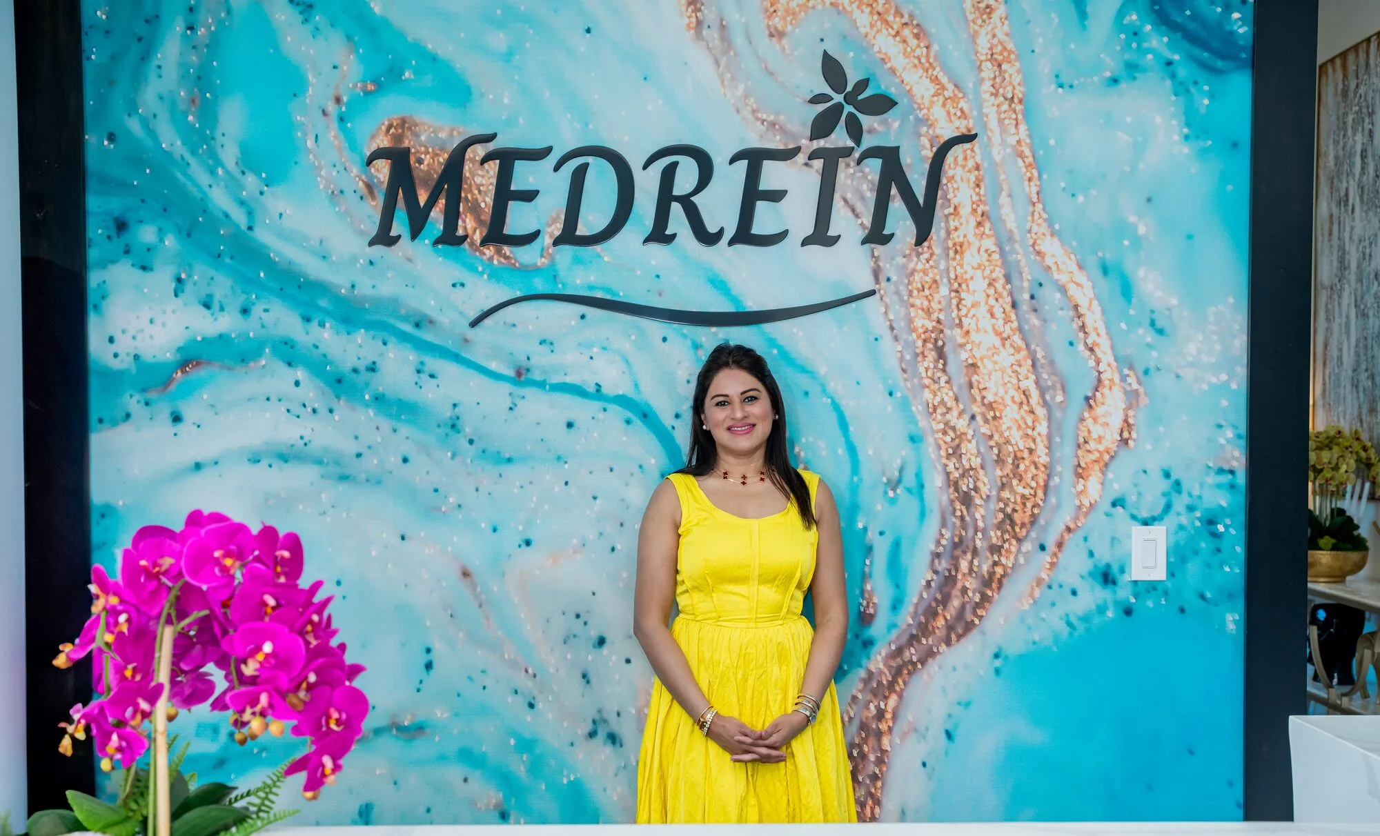 Dr. Haritha Mikkilineni, MD, standing at the reception desk beneath the MEDREIN mural at the Medrein Health clinic in Southlake, TX