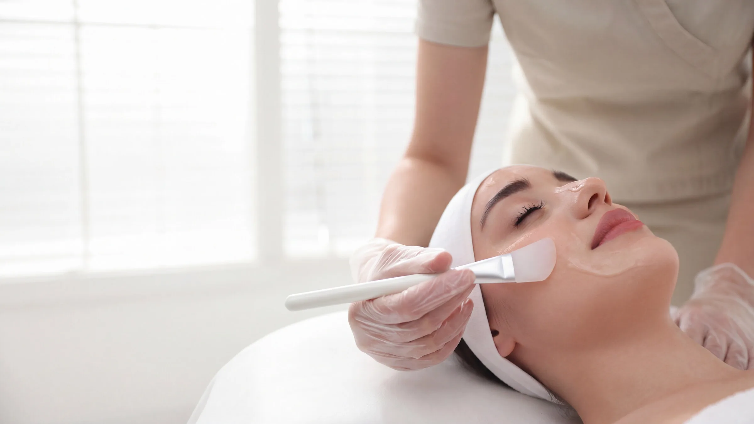 Wide view of a medical-grade facial in progress, a gloved clinician brushing treatment onto a reclining patient's skin in a bright, daylit treatment room