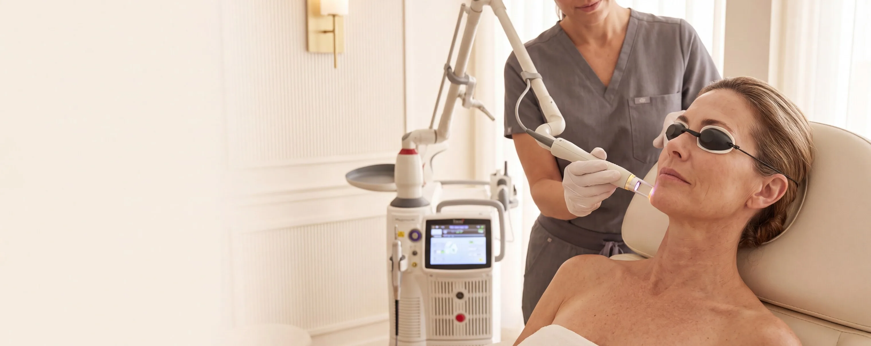 A patient in protective eye shields reclined in a treatment room while a clinician guides a laser handpiece along her cheek, the Fotona SP Dynamis dual-wavelength console and its articulated arm standing behind them