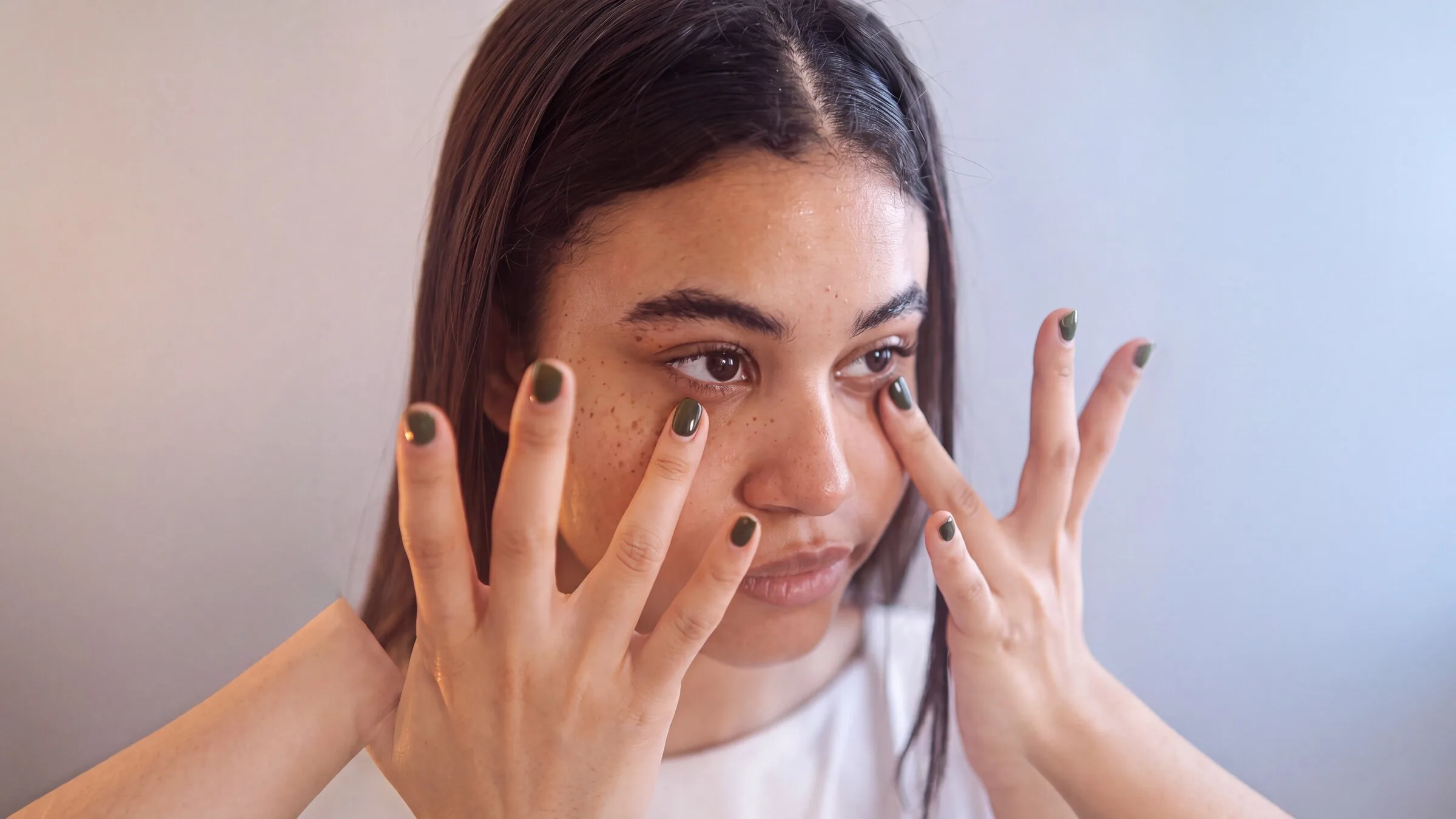 A woman resting a fingertip below each eye, soft brown-violet shadowing under both lower lids, against a plain out-of-focus wall