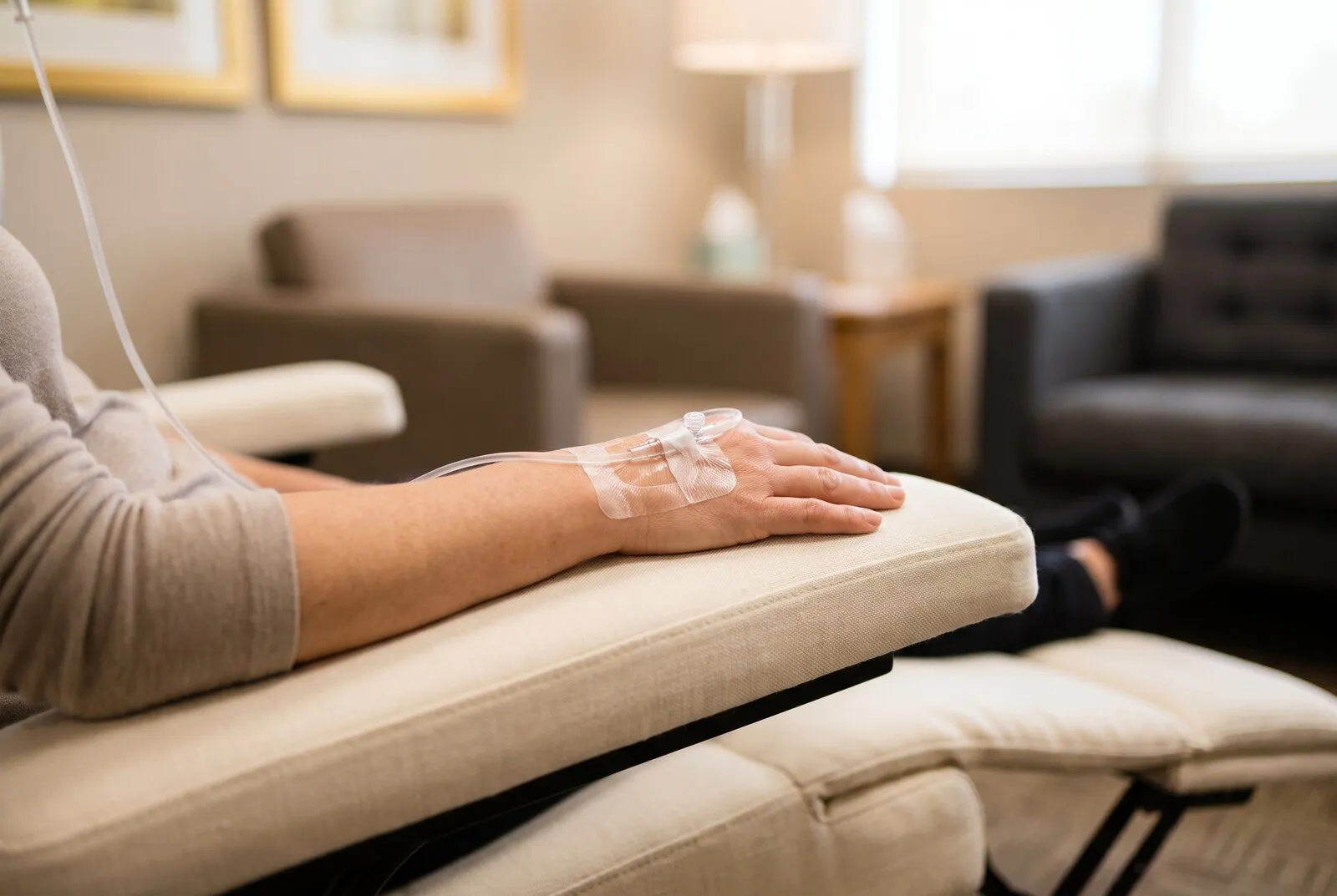 A patient’s forearm resting on the cream armrest of a reclining clinic chair, an intravenous catheter taped to the back of the hand with a clear infusion line running out of frame