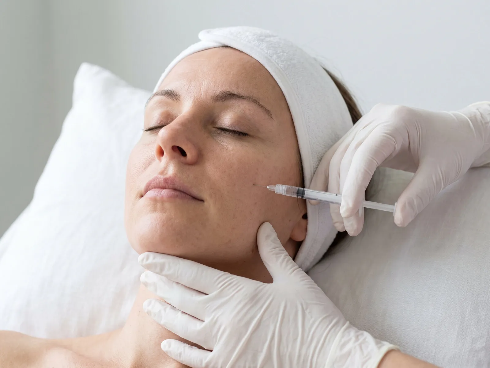 Close view of a gloved clinician placing a fine dermal filler needle at the mid-cheek of a reclining patient