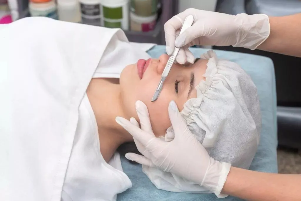 A patient lying on a treatment couch in a clinic while a gloved aesthetician holds a sterile blade against her forehead