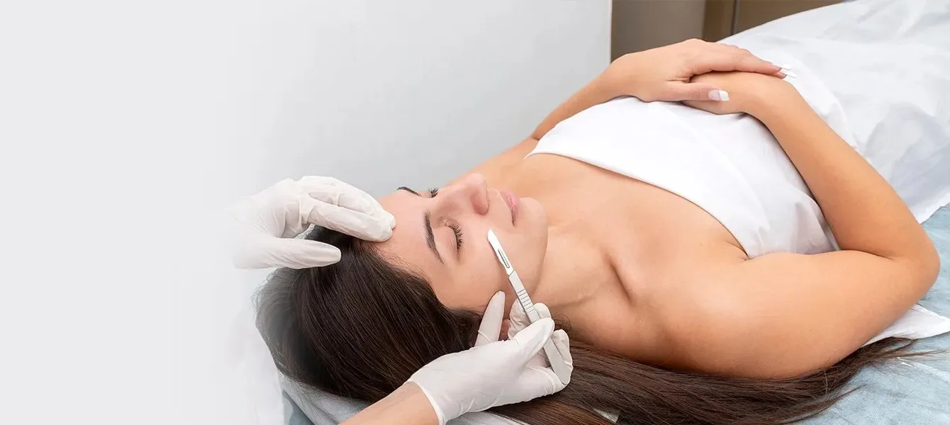 A patient reclining on a treatment couch while a gloved clinician draws a sterile dermaplaning blade across her cheek