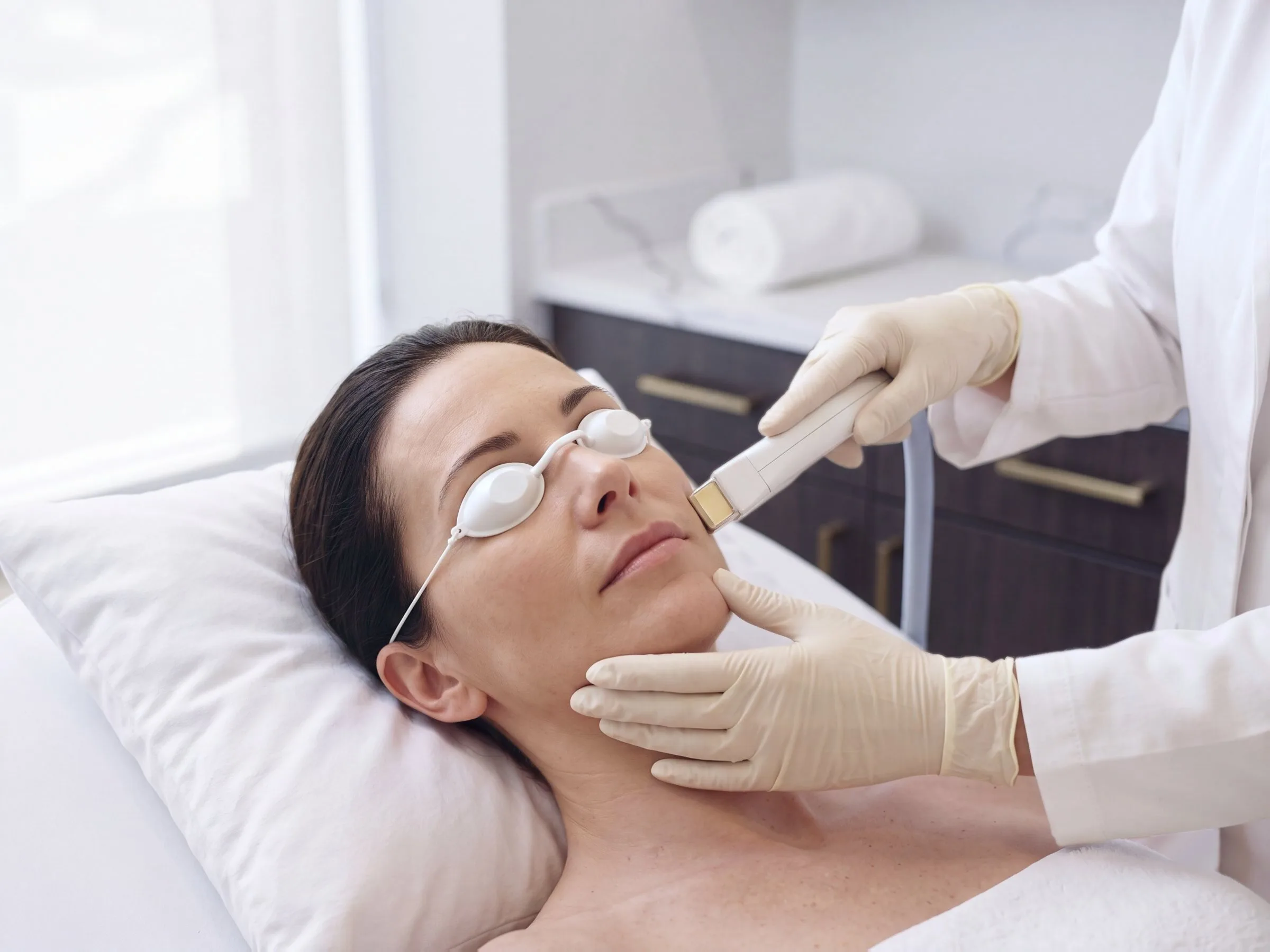 A clinician passing a laser handpiece along a reclining patient's cheek and jawline during a Fotona 4D facelift session in a cool white treatment room with espresso cabinetry at Medrein Health in Southlake, TX