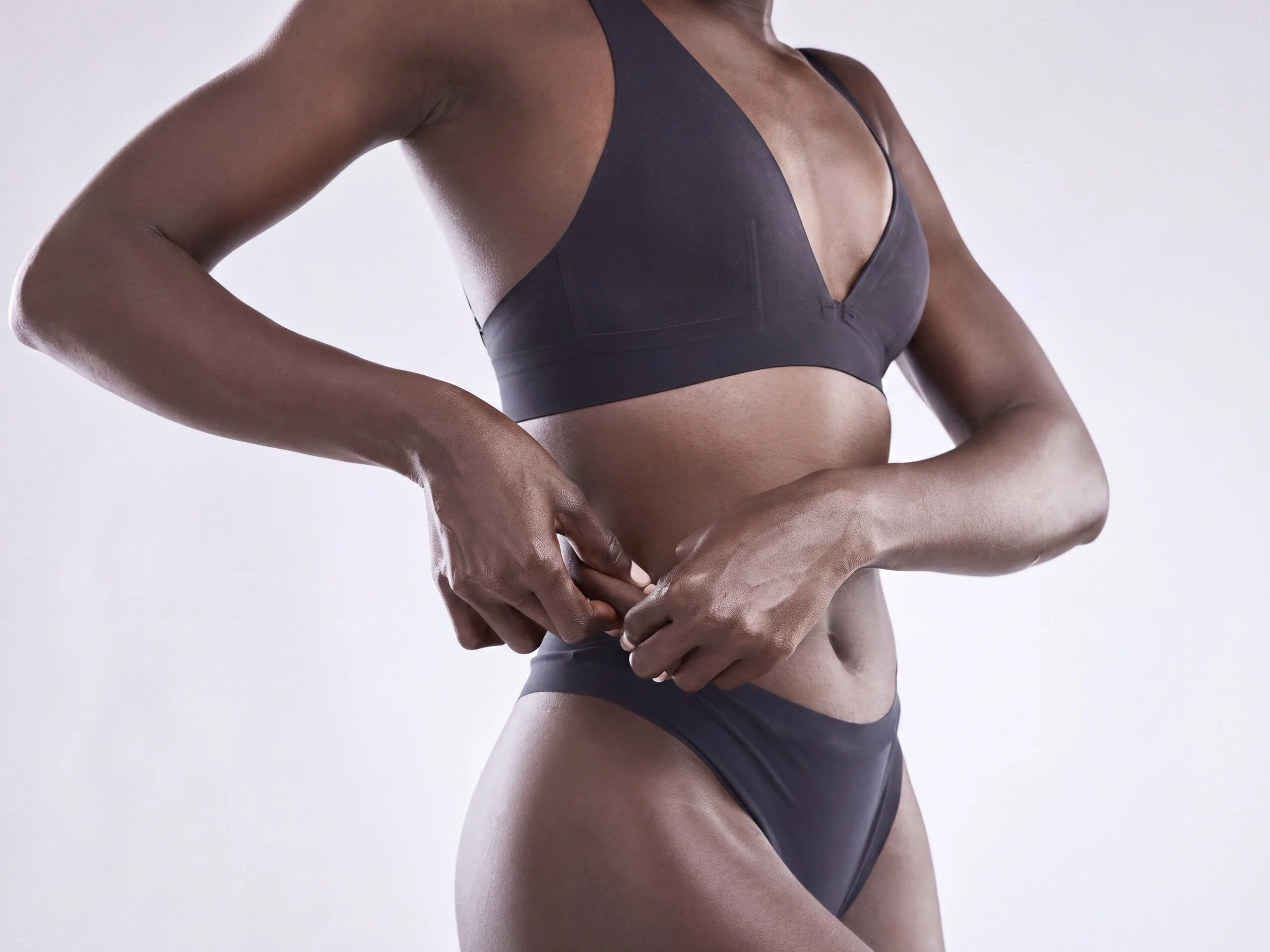 A woman in a charcoal bra and briefs standing against a pale grey studio background, both hands resting at her waist