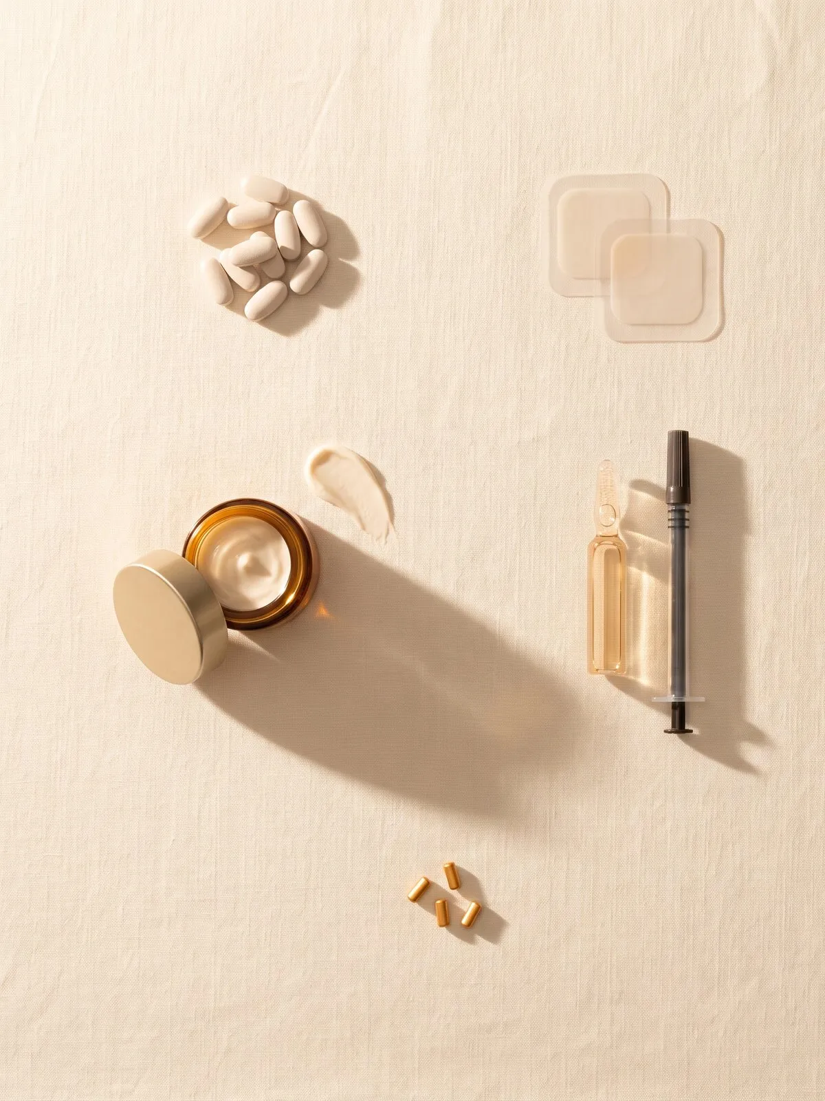 Overhead view on cream linen of the five forms bioidentical hormones come in: a pile of pale tablets, two square transdermal patches, an open amber jar of cream with a swipe beside it, a glass ampoule with a syringe, and four small gold pellets