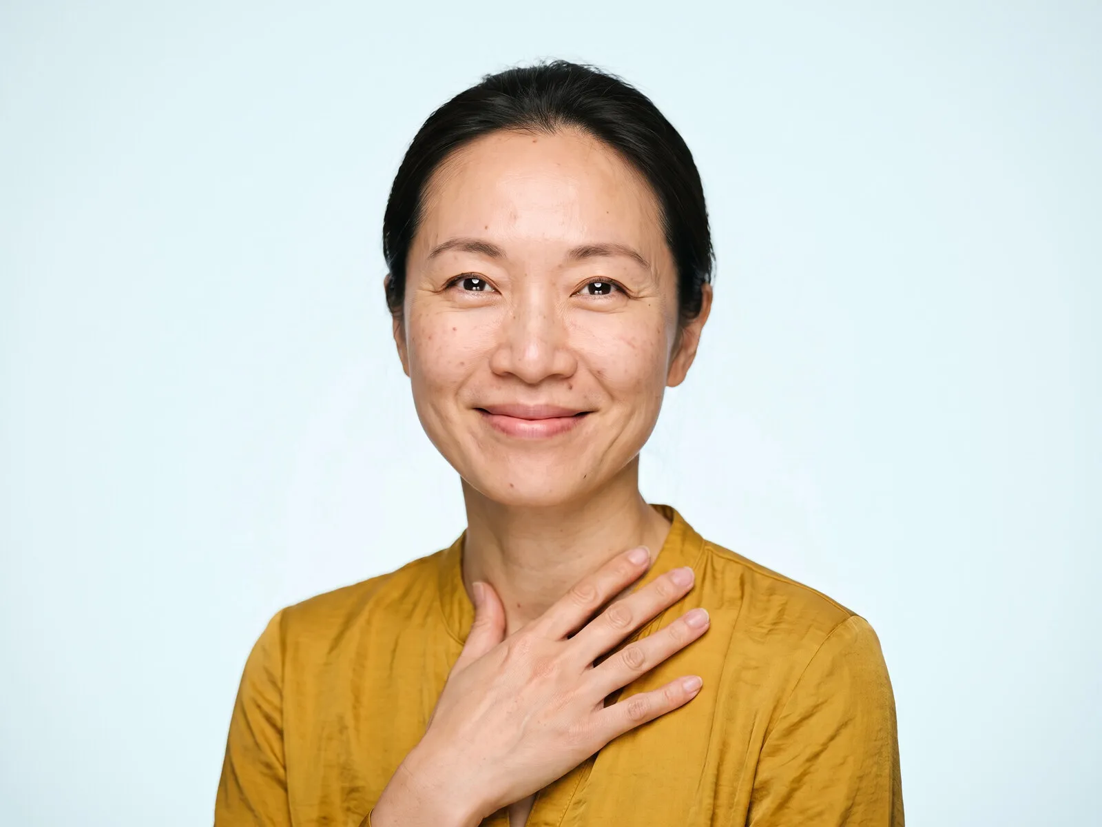 A woman in her mid forties with a hand resting at her collarbone, smiling to camera