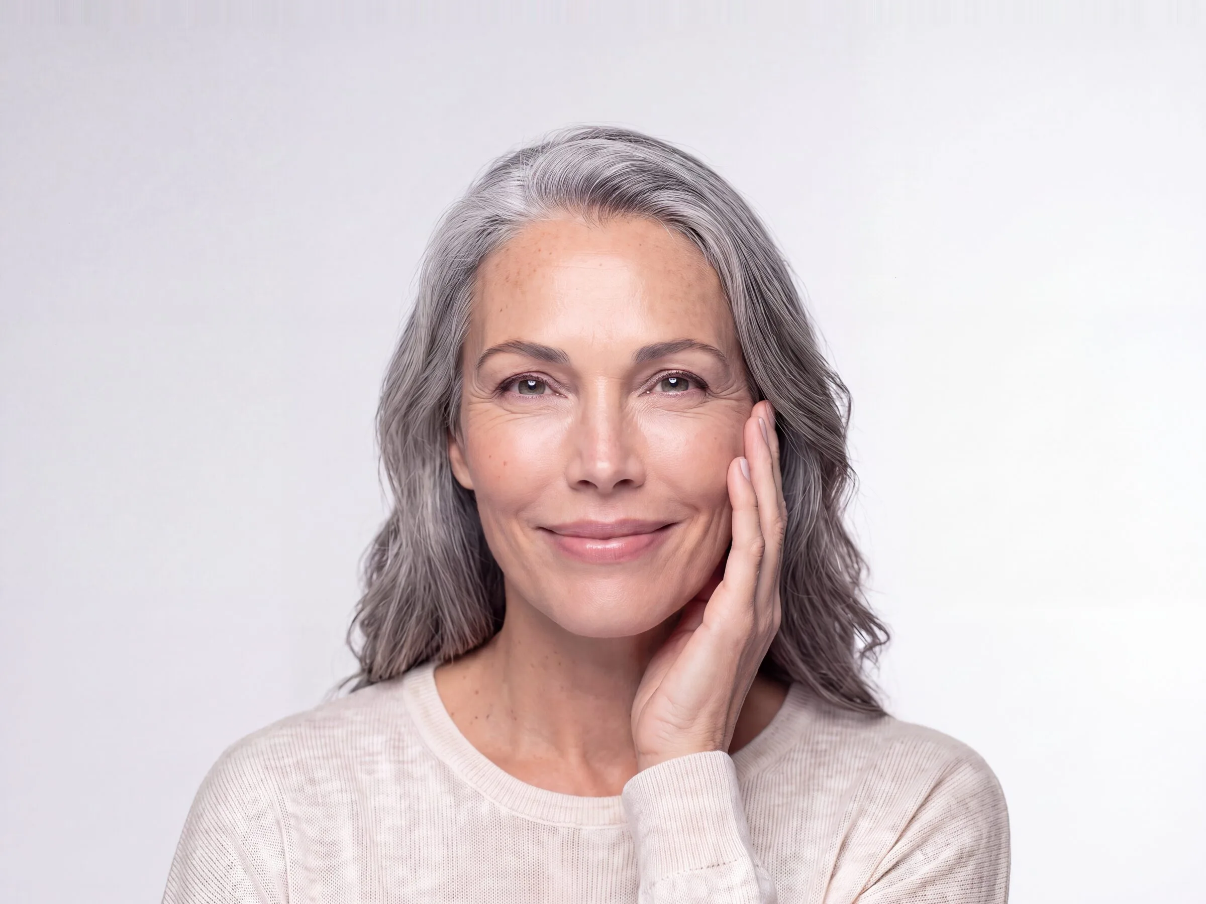 Close-up of a woman in her fifties with long silver-grey hair resting her fingertips lightly against her cheek, smiling gently to camera in soft studio light against a plain pale grey background