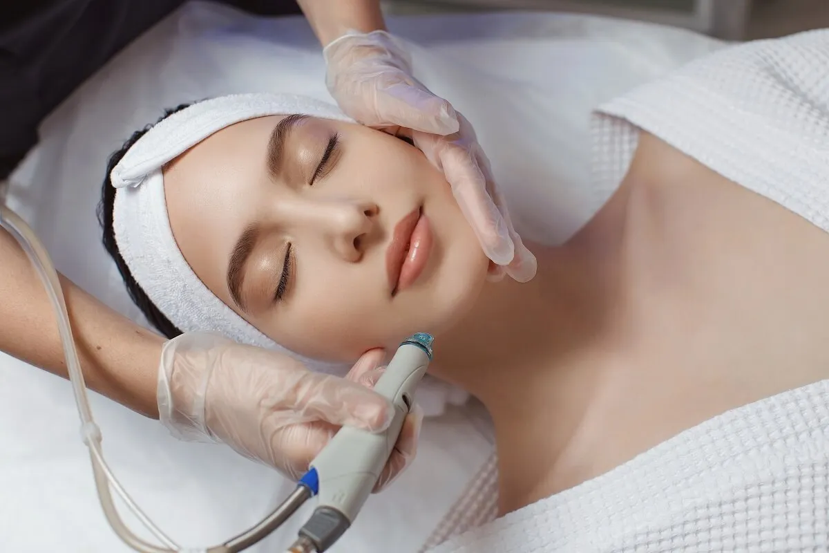 Gloved clinician holding a hydradermabrasion handpiece against the jawline of a reclining patient in a white headband during a facial