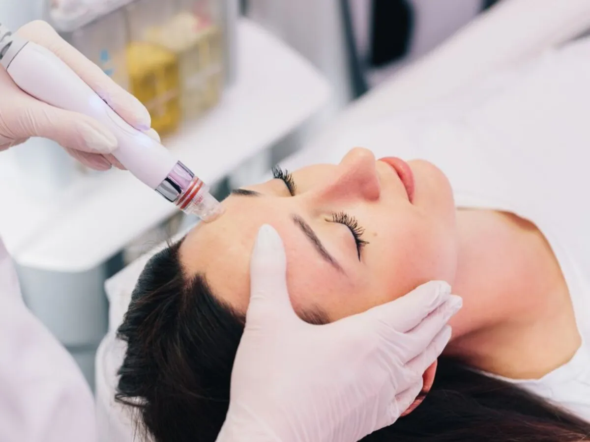 Gloved clinician holding a hydradermabrasion handpiece to a patient’s cheek beside the HydraFacial machine