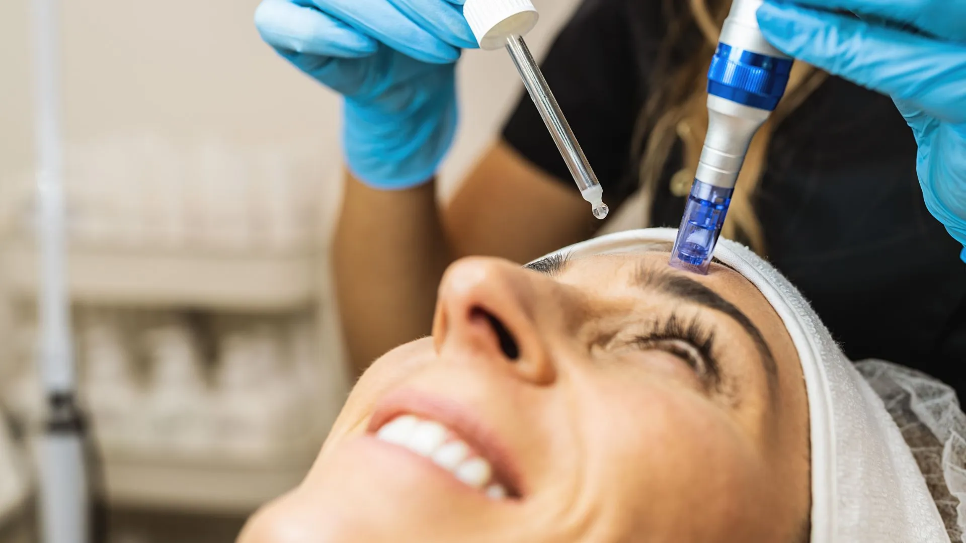 A gloved clinician drawing serum into a dropper while working a microneedling handpiece across a smiling patient’s forehead