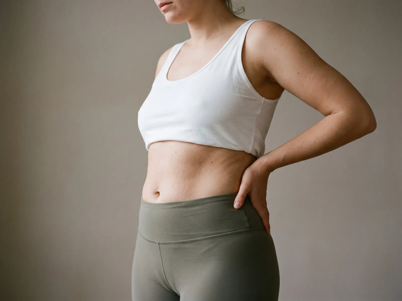 A woman standing with a hand on her waist in a cropped white top and olive leggings, showing the untouched midsection and waist clients ask about when tone rather than weight is the goal