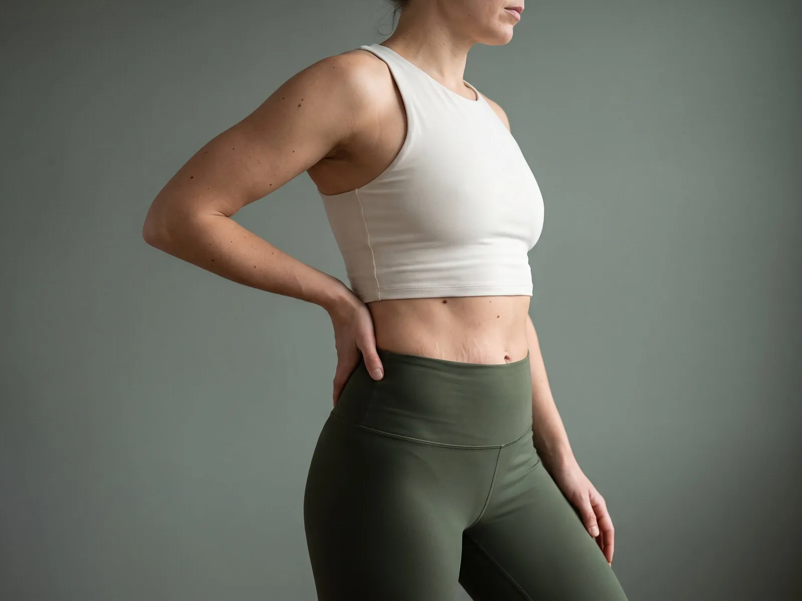 A woman standing turned three-quarters away in an off-white cropped top and olive leggings with a hand at her hip, the abdomen and core that an MNML Tone session builds tone in