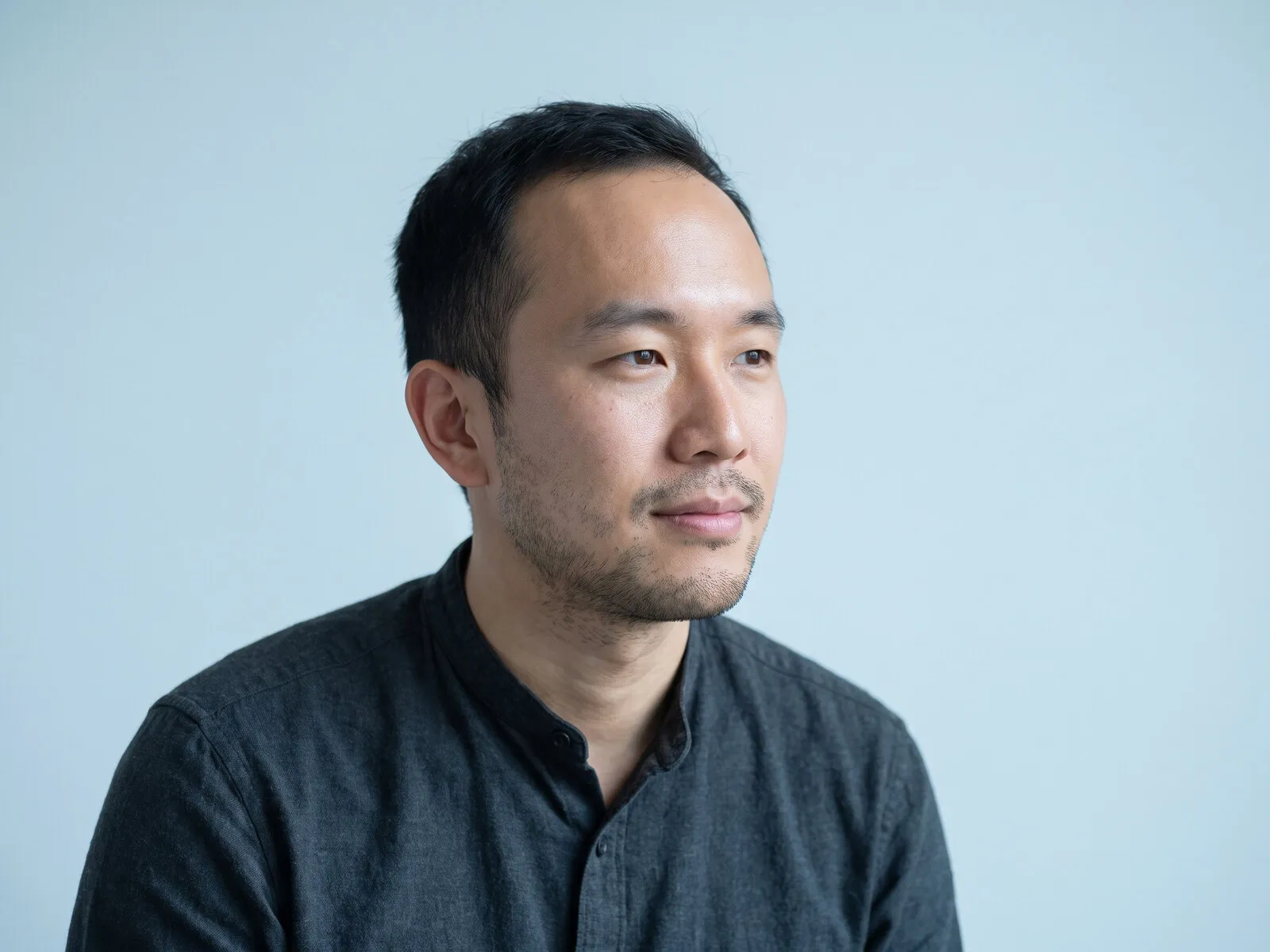 A man in his early thirties in close portrait, turned three-quarters and looking off-camera in concentrated attention, on a pale azure ground