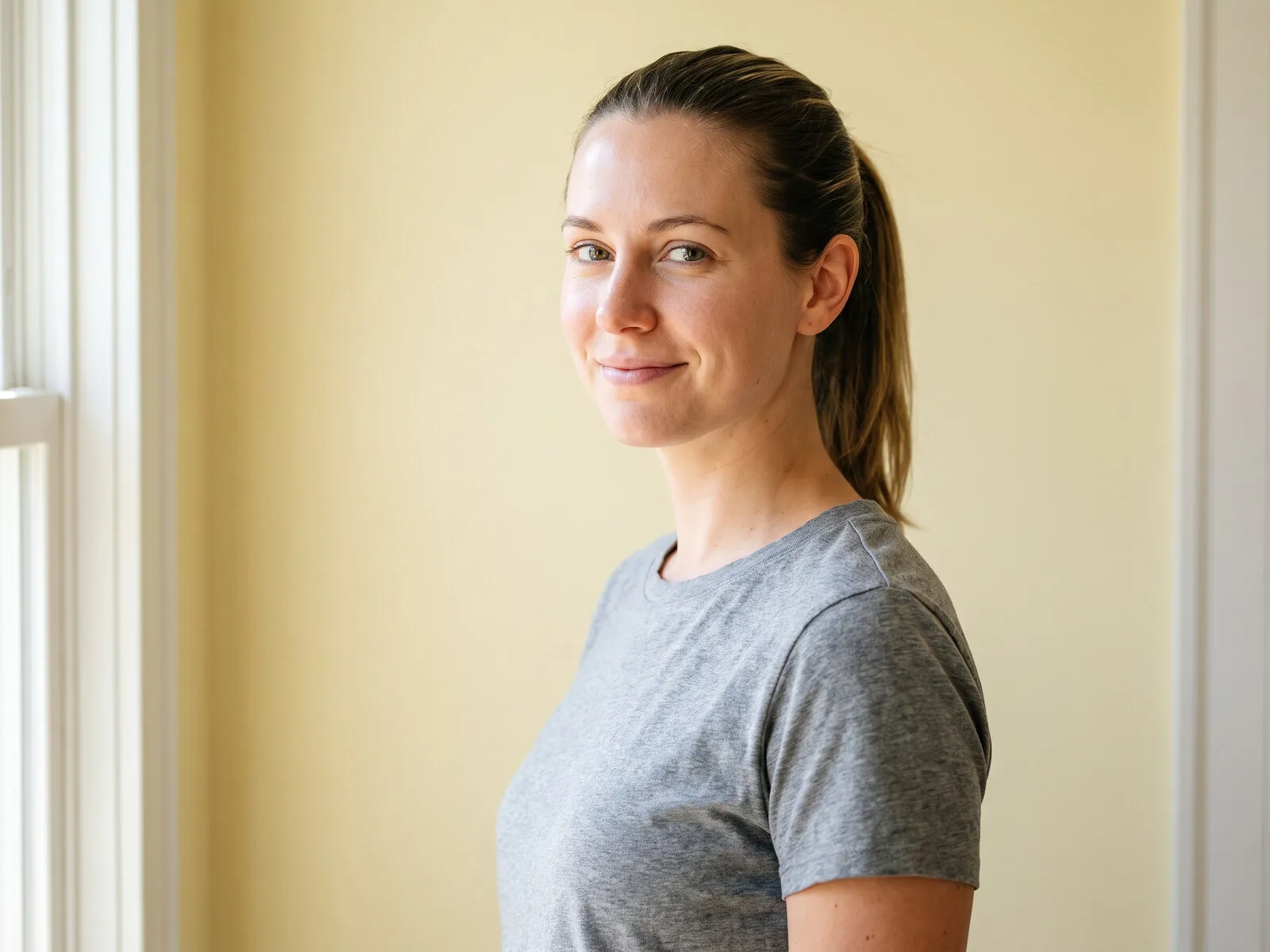 A woman in her late twenties standing beside a window in daylight, turned to the camera with an easy smile, against a soft yellow wall