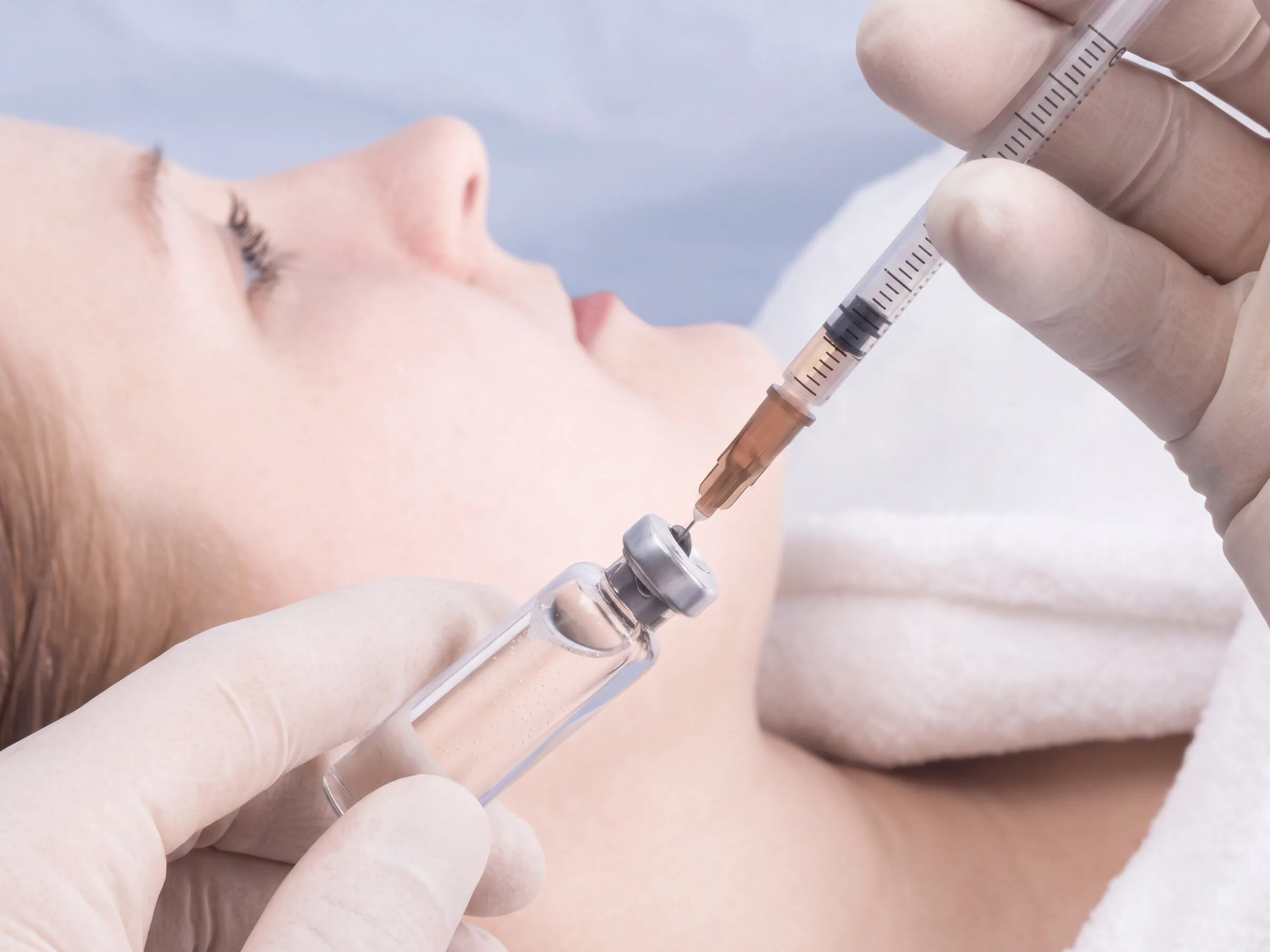 A gloved clinician drawing a prescription peptide from a small glass ampoule into a graduated syringe, a patient resting on a treatment table behind