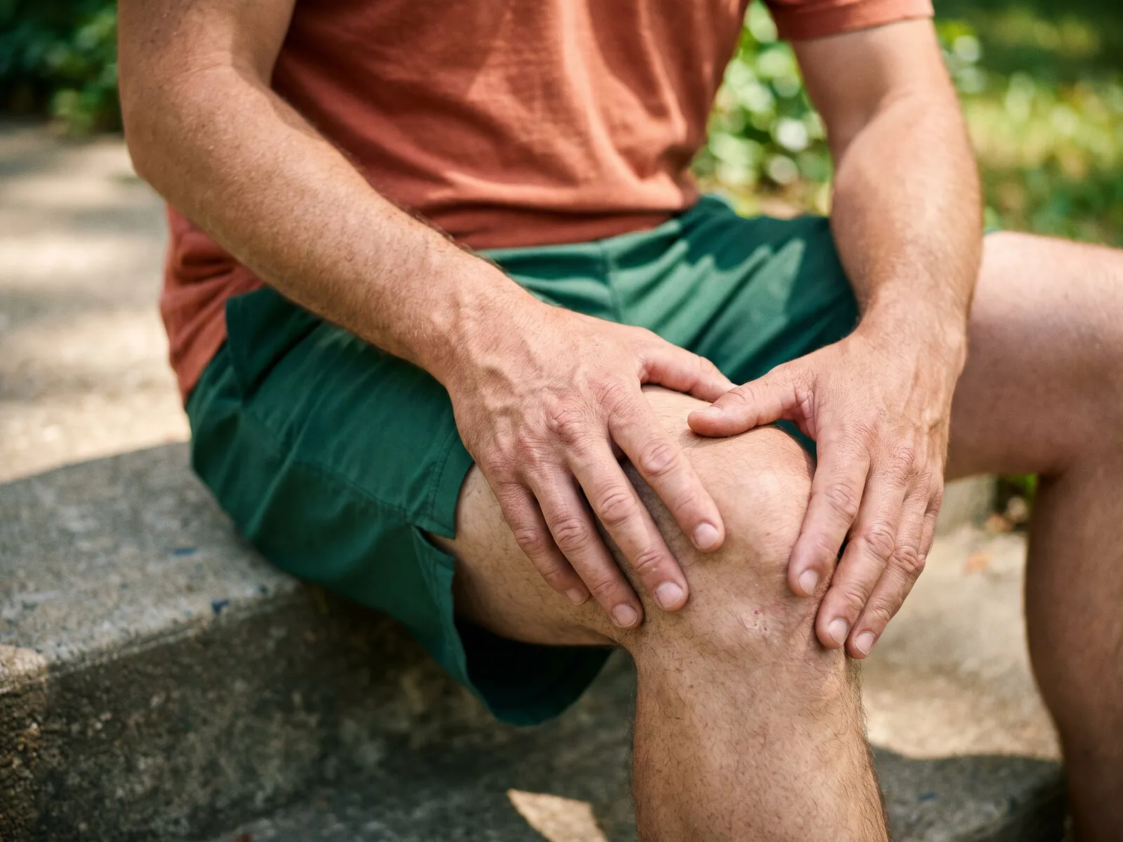 A man in running clothes sitting on a low wall outdoors with both hands resting on a sore knee