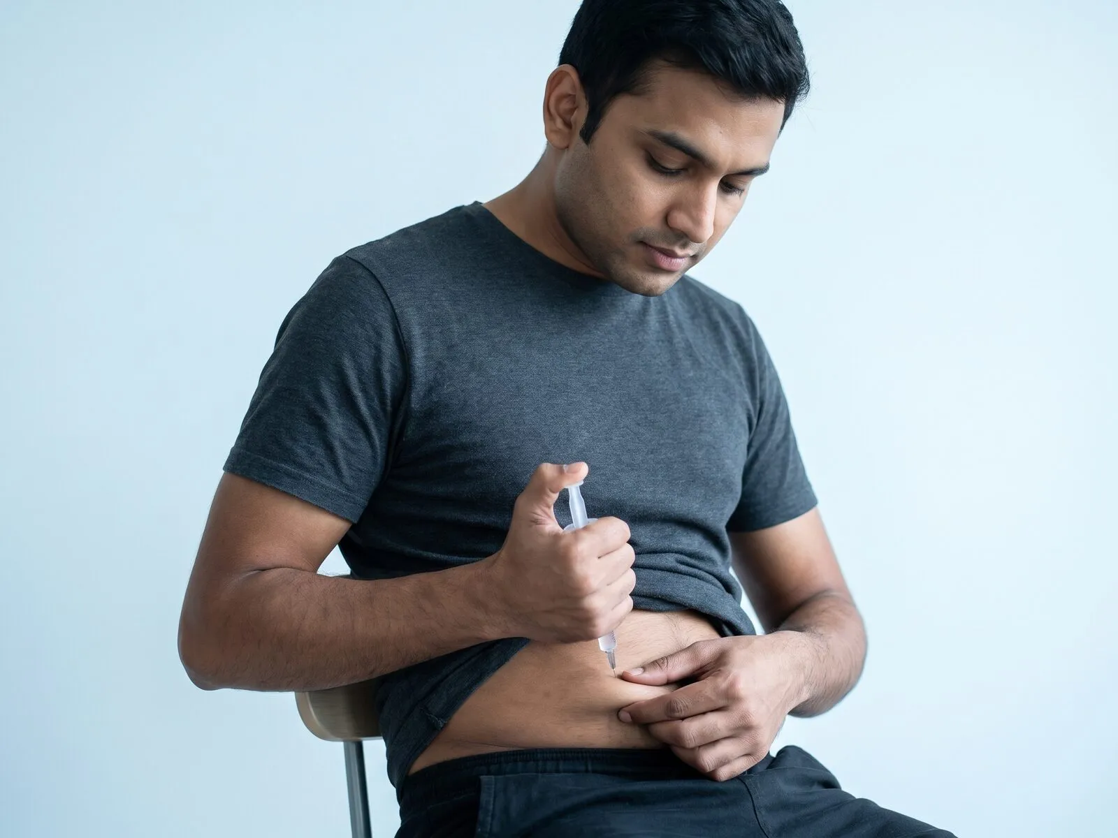 A man in his late twenties seated against a pale blue studio backdrop, giving himself an evening subcutaneous injection in a pinched fold of his lower abdomen with a small syringe