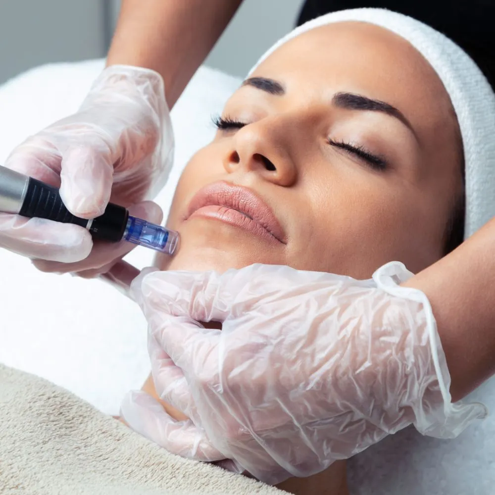 A gloved clinician guiding a microneedling handpiece along the jawline of a patient reclining on a treatment bed